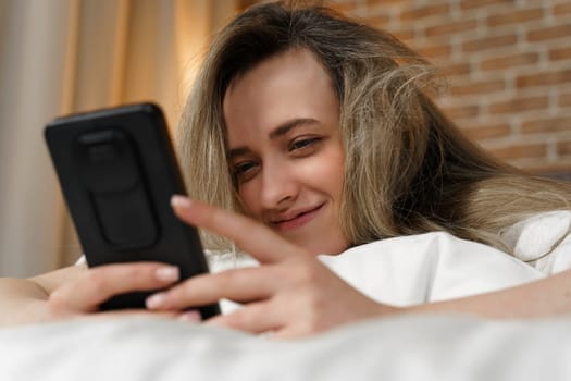 A young beautiful woman is lying on the bed, watching content in a smartphone, smiling. Close-up.