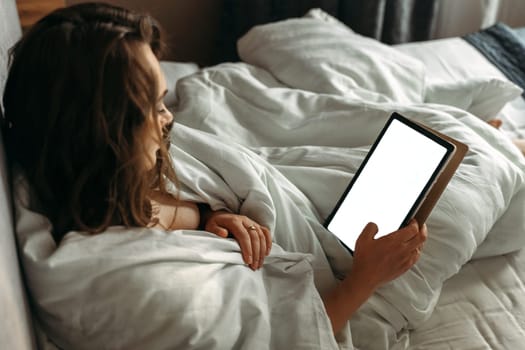 A young woman lies in bed in the morning, looks at the tablet. Mockup