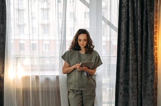 A young woman in a suit stands at the window with a tablet in her hands. Browses online stores, social networks.