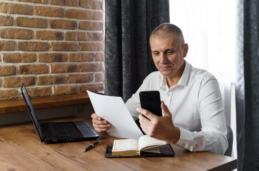 A businessman holds a phone and documents in his hands, reads information from the phone. Financial manager, company.