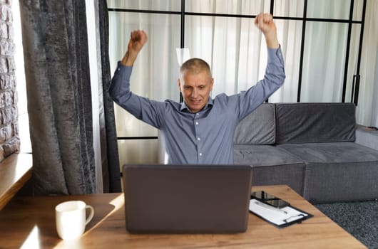 A businessman working at a computer rejoices in a perfect successful transaction, raising his hands up. The concept of success.