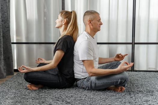 A man and a woman sit back to back, practice yoga and meditate for relaxation and balance of life in a room at home. Sports concept.