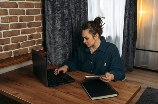 A beautiful woman sits at a table indoors and works remotely on a tablet and laptop.