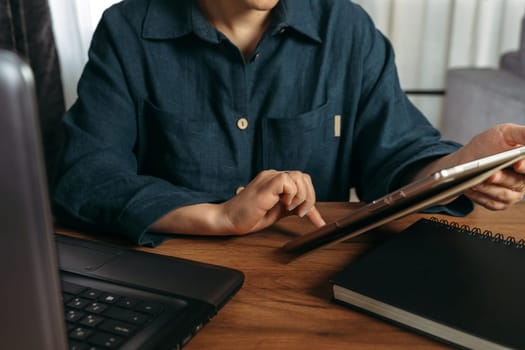 A beautiful woman sits at a table indoors and works remotely on a tablet. Close-up.