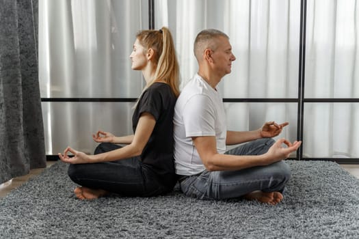 A man and a woman sit back to back, practice yoga and meditate for relaxation and balance of life in a room at home. Sports concept.
