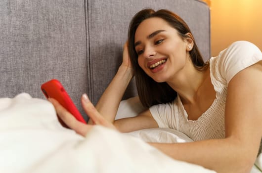 A beautiful young girl lies in bed and enjoys communication, holds a smartphone in her hands.