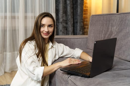 A beautiful young female freelancer is working on a laptop which is standing on the sofa, she is sitting on the floor herself.