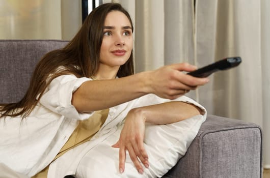 Positive young woman sitting on the sofa indoors at home and watching TV with remote control on the sofa.