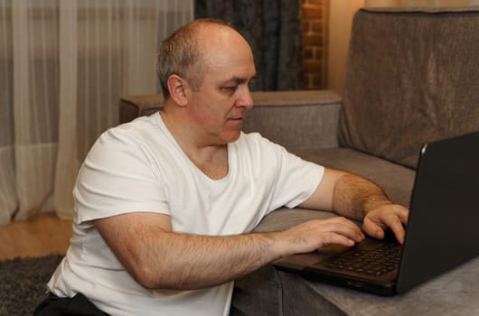 A mature man sits on the floor and works on a laptop that is on the sofa.