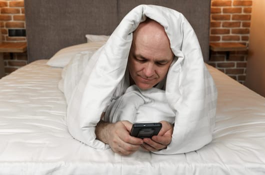 An adult man lies covered with a blanket and browses social networks, videos in a smartphone.