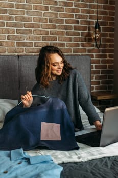 A young woman sits on a bed and admires her purchases made through an online store. Writes a review about the purchase of goods on a laptop.