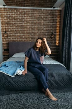 A beautiful woman in designer clothes sits on a bed, clothes lie nearby. Vertical frame.