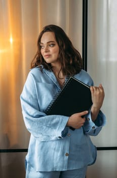 A portrait of a young woman in a light blue suit is standing in a room with a tablet and a notebook in her hands. Vertical frame.