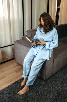 A young woman in a blue suit sits on the back of a sofa with a tablet in her hands. Browses online stores, social networks. Vertical frame.