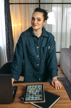 Portrait of a business woman standing near the desk. Notepad, tablet and laptop on the table. Vertical frame.