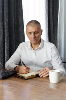 A middle-aged man sits at a table, works, writes down information in a notebook. Vertical frame. Remote work at home.