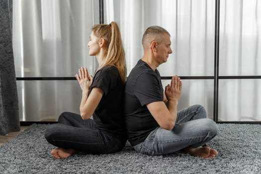 A man and a woman sit back to back, practice yoga and meditate for relaxation and balance of life in a room at home. Sports concept.