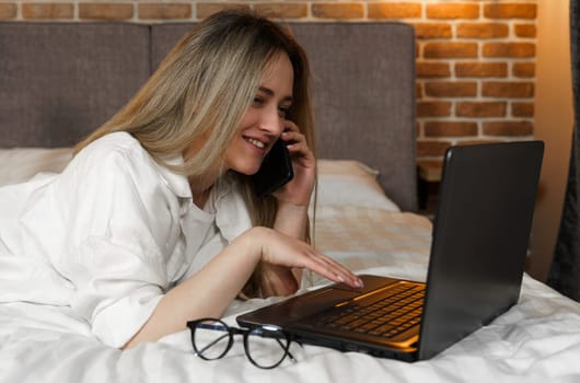 A young beautiful woman is lying on the bed, talking on the phone, looking into a laptop.