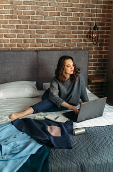 Women's outfits are laid out on the bed, a woman is sitting next to the bed and working at the computer. Vertical frame.