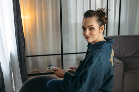 A young woman in a dark blue suit sits on the back of a sofa with a tablet in her hands. Browses online stores, social networks. Back view.