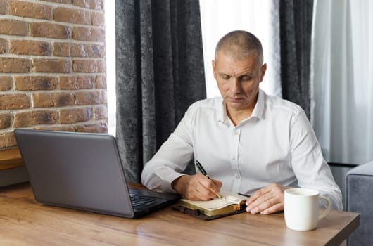A middle-aged man sits at a table, works, writes down information in a notebook. Remote work at home.