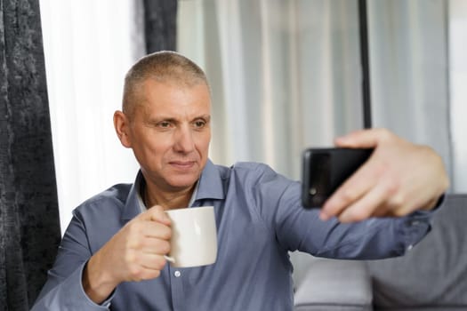 A middle-aged man with tea takes a selfie photo with a mobile phone in the apartment.