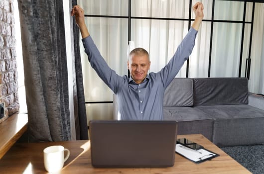 A businessman working at a computer rejoices in a perfect successful transaction, raising his hands up. The concept of success.