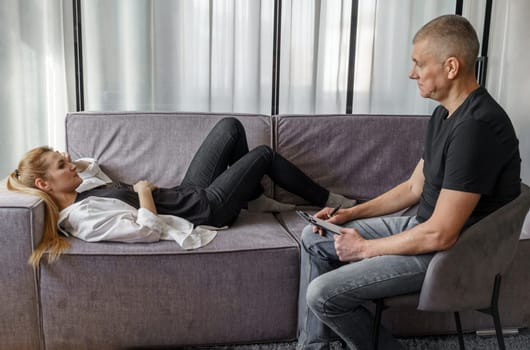 A male psychologist sits on a chair and talks to a patient in a psychotherapy session. Positive adult woman with mental health problems lies on the couch and communicates with a psychologist.