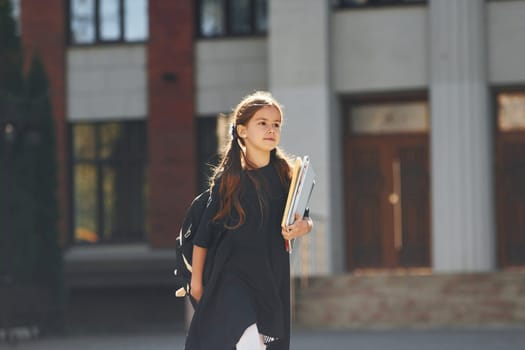 Schoolgirl is walking outside near school building.