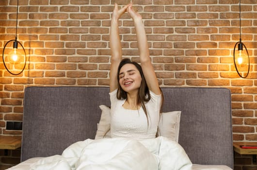 Beautiful woman sits on the bed, stretches her arms up after sleeping.