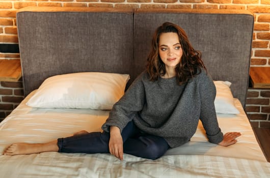 An attractive woman is sitting on a bed in a bedroom wearing blue trousers and a gray jumper. Smiling and looking away.