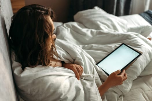 A young woman lies in bed in the morning, watches the news on her tablet.