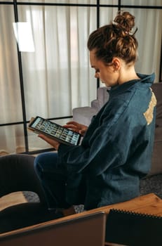 A young woman in a dark blue suit sits on the back of a sofa with a tablet in her hands. Browses online stores, social networks. Back view. Vertical frame.