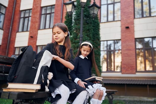 Sitting together. Two schoolgirls is outside near school building.