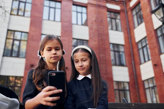 Sitting together. Two schoolgirls is outside near school building.