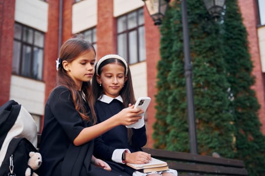 Sitting together. Two schoolgirls is outside near school building.
