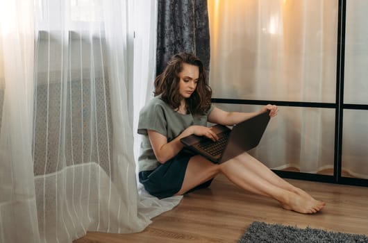 A beautiful woman is sitting on the floor near the window with a laptop, working or studying.