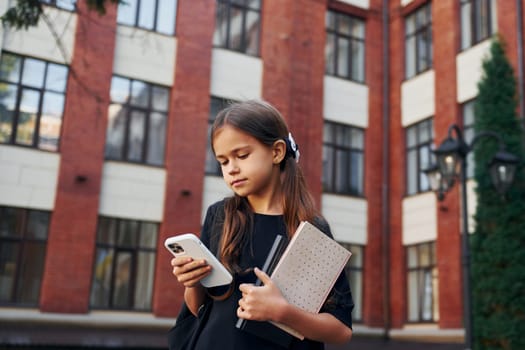 Holding phone in hands. Schoolgirl is outside near school building.