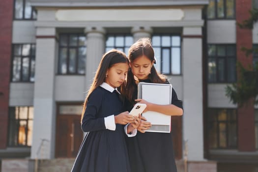 Front view. Two schoolgirls is outside together near school building.