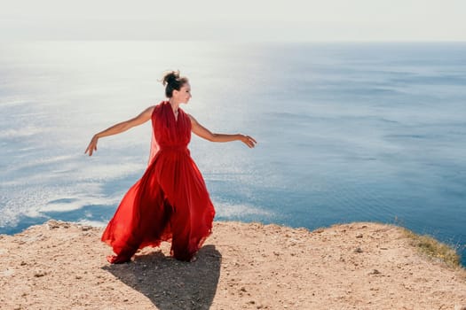 Side view a Young beautiful sensual woman in a red long dress posing on a rock high above the sea during sunrise. Girl on the nature on blue sky background. Fashion photo.