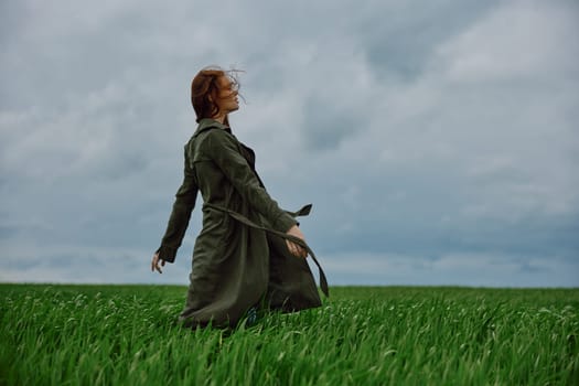 a red-haired woman in a long raincoat stands in a green field in cloudy weather in a strong wind. High quality photo