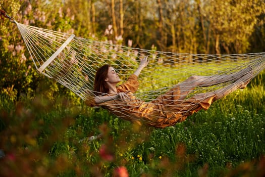 a beautiful woman is resting in nature lying in a mesh hammock in a long orange dress looking to the side, arms outstretched. Horizontal photo on the theme of recreation. High quality photo