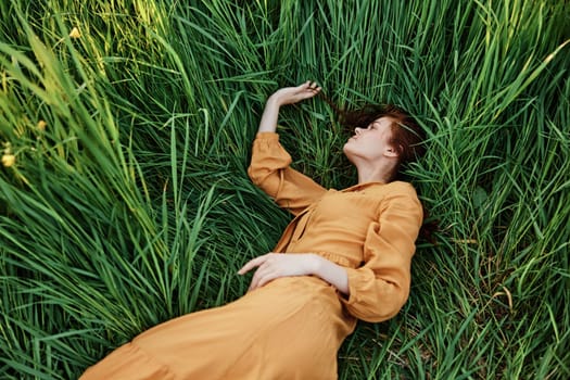 a close horizontal photo of a pleasant woman in a long orange dress resting lying in the tall grass with her eyes closed in sunny weather. High quality photo