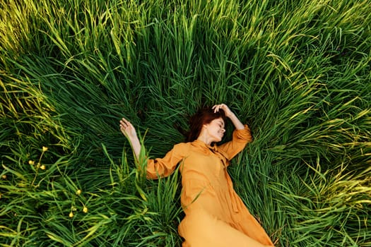 an elegant woman lies in the tall green grass in a long summer orange dress and with her arms outstretched enjoys nature and sunny weather with her eyes closed. High quality photo