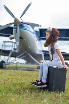 A little girl in a pilot's costume sits on a suitcase against the background of an airplane with a propeller. Child Dreams of flying in the sky.