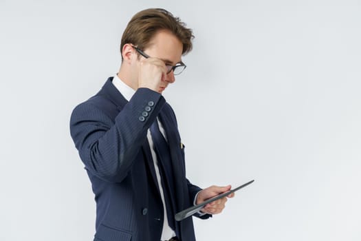 Portrait of a businessman in a blue suit who is holding an electronic tablet in his hands and adjusting his glasses. White background. Business and finance concept