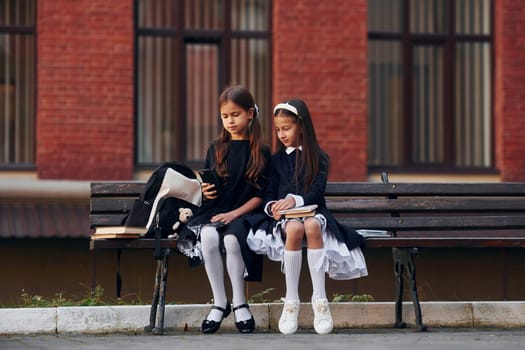 Two schoolgirls is sitting outside together near school building.