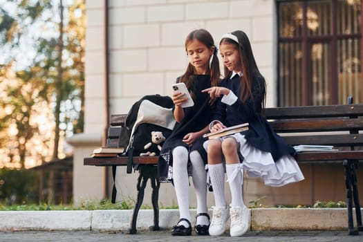 Two schoolgirls is sitting outside together near school building.