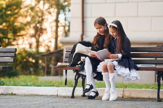 Two schoolgirls is sitting outside together near school building.