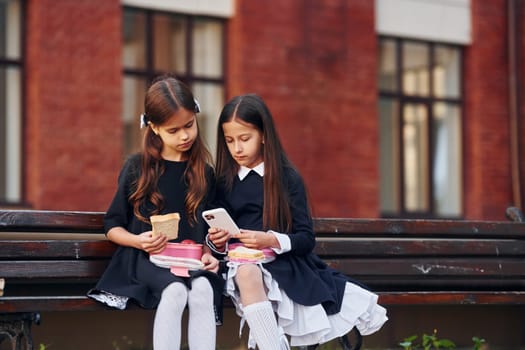 Two schoolgirls is sitting outside together near school building.